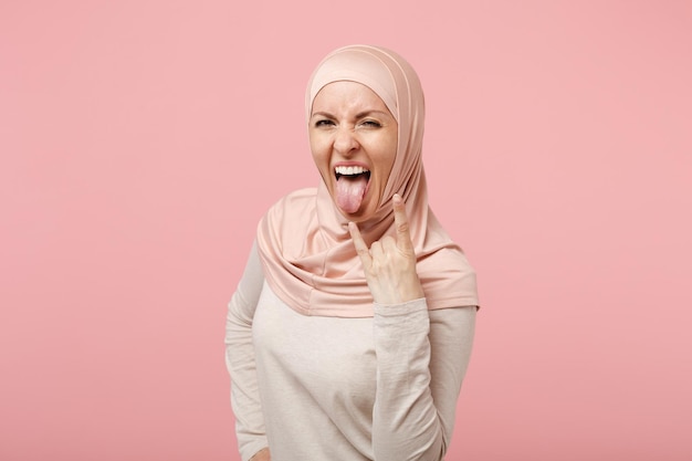 Donna musulmana araba in abiti leggeri hijab in posa isolata su sfondo rosa. Concetto di stile di vita religioso della gente. Mock up spazio di copia. Mostra la lingua raffigurante un segno di metallo pesante con le corna in alto.