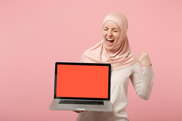 Donna musulmana araba in abiti leggeri hijab in posa isolata su sfondo rosa. Concetto di stile di vita religioso della gente. Mock up spazio di copia. Tenere il computer portatile con schermo vuoto, facendo il gesto del vincitore.