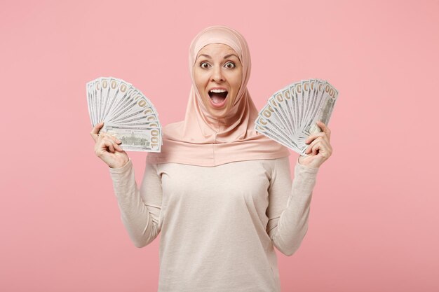 Donna musulmana araba sorpresa in abiti leggeri hijab in posa isolata su sfondo rosa. Concetto di stile di vita dell'Islam religioso della gente. Mock up spazio di copia. Tenendo fan di denaro contante in banconote in dollari.