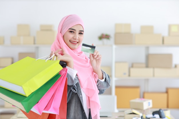 Donna musulmana asiatica astuta attiva nella condizione del rivestimento di jeans e nei sacchetti della spesa della tenuta mentre mostrando la carta di credito con il fondo online di consegna della scatola del pacchetto. Bella ragazza che esamina cemera e sorriso.