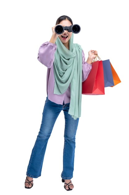 Donna musulmana asiatica in un velo che guarda attraverso il binocolo mentre tiene le borse della spesa