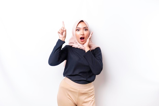 Donna musulmana asiatica scioccata che indossa l'hijab che punta allo spazio della copia sopra di lei isolato da sfondo bianco