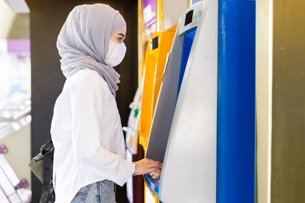 Donna musulmana che indossa una maschera medica per prevenire il virus dell'infezione usando il bancomat per prelevare denaro sulla strada della città.