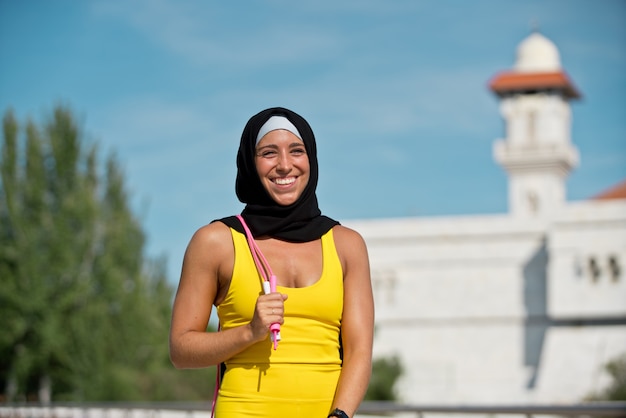 Donna musulmana fitness che indossa l'hijab in abbigliamento sportivo giallo isolato con corda per saltare. Vista laterale orizzontale della donna araba che si allena all'aperto con sfondo moschea.