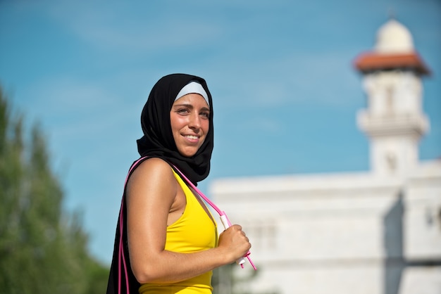 Donna musulmana fitness che indossa l'hijab in abbigliamento sportivo giallo isolato con corda per saltare. Vista laterale orizzontale della donna araba che si allena all'aperto con sfondo moschea.