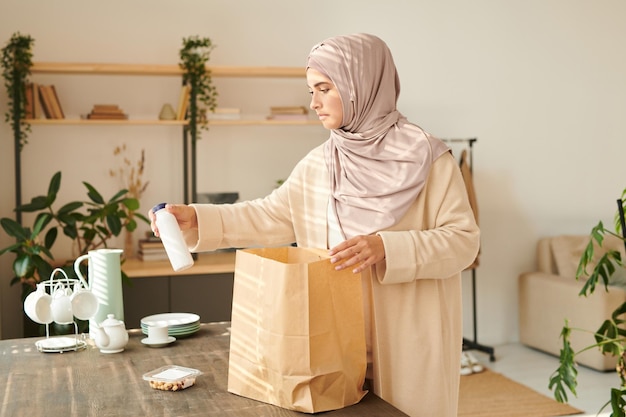 Donna musulmana moderna in foulard in piedi a tavola che tira fuori i prodotti alimentari dal sacchetto di carta che le è stato consegnato