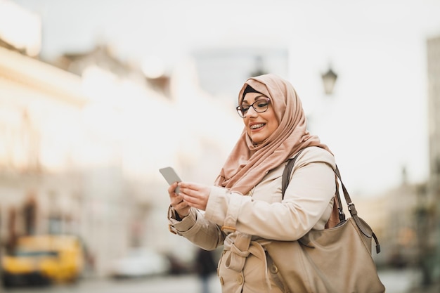 Donna musulmana sorridente che indossa un hijab e usa il suo smartphone mentre si trova in un ambiente urbano.