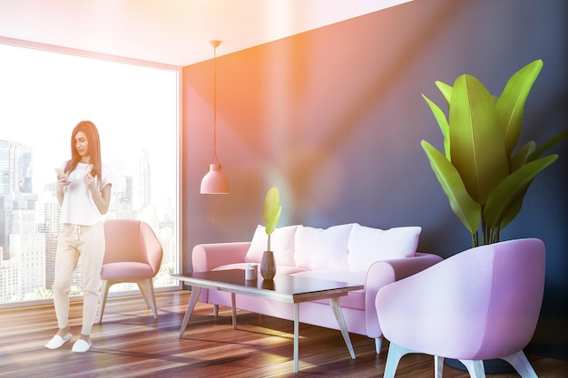 Donna nell'angolo del soggiorno panoramico con pareti blu, divano rosa e due poltrone rosa in piedi vicino al tavolino nero. Immagine tonica