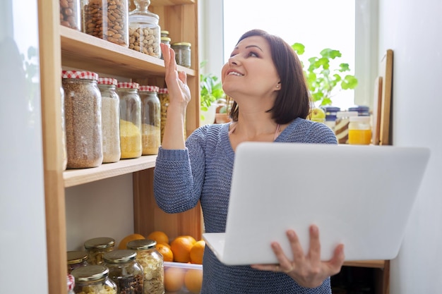 Donna nella dispensa della cucina con i prodotti immagazzinati che tengono il computer portatile