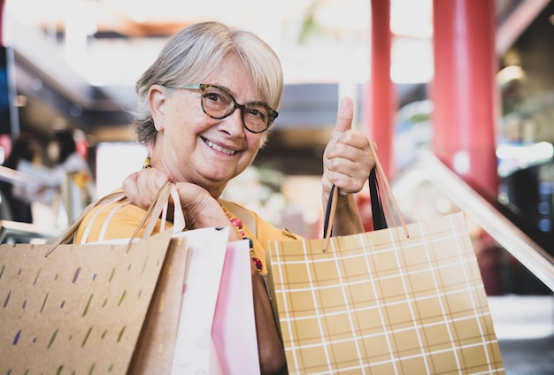 Donna nello shopping. Felice donna anziana con borse della spesa sulla scala mobile del centro commerciale, con il pollice in alto come concetto positivo ottimista. Anziano giovane con gli occhiali