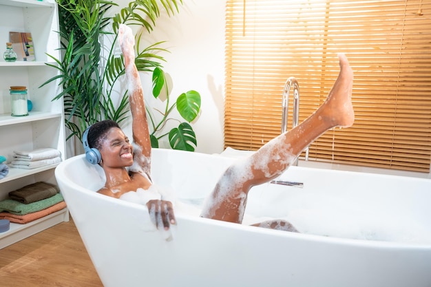 Donna nera attraente che indossa le cuffie con la musica nella vasca da bagno di schiuma