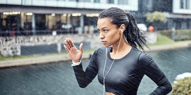 Donna nera fitness e corsa con auricolari sotto la pioggia per motivazione sportiva o determinazione in città Corridore sportivo afroamericano che fa un intenso allenamento cardio sotto la pioggia