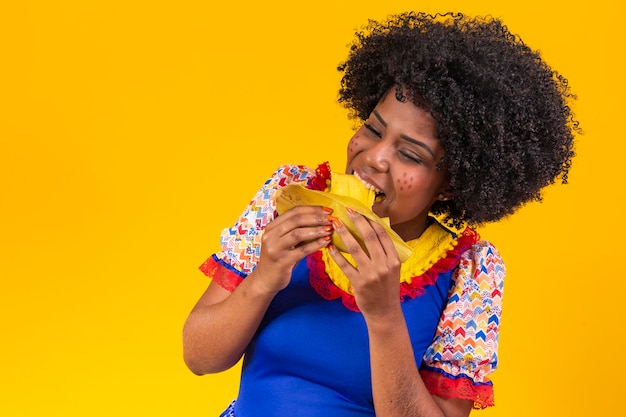 Donna nera in abiti tipici festa junina che mangiano deliziosi pamonha su sfondo giallo spazio libero per il testo