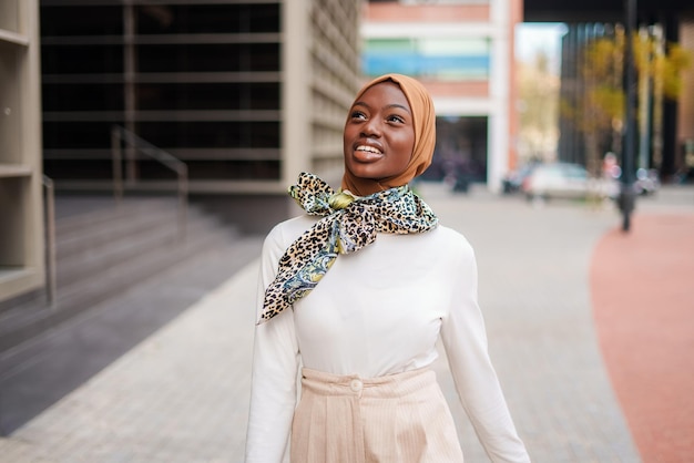 Donna nera musulmana sorridente in hijab in piedi vicino all'edificio per uffici