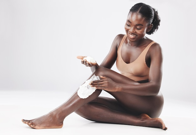 Donna nera seduta con crema da barba sulle gambe per cosmetici per la cura della pelle e benessere della pelle con il sorriso Donna africana felice che cura i peli del corpo e cura di sé amore e salute sullo sfondo dello studio