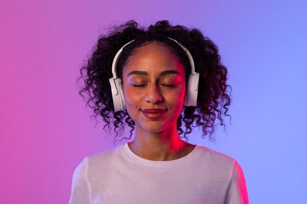 Donna nera soddisfatta con le cuffie che si diverte a ascoltare musica sullo sfondo al neon