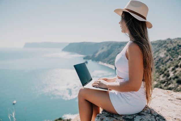 Donna nomade digitale nel cappello una donna d'affari con un laptop si siede sulle rocce in riva al mare durante