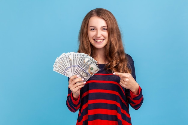 Donna ottimista che indossa un maglione a righe in stile casual che punta il dito e un grande fan di banconote in dollari guardando la fotocamera con espressione soddisfatta Colpo in studio al coperto isolato su sfondo blu
