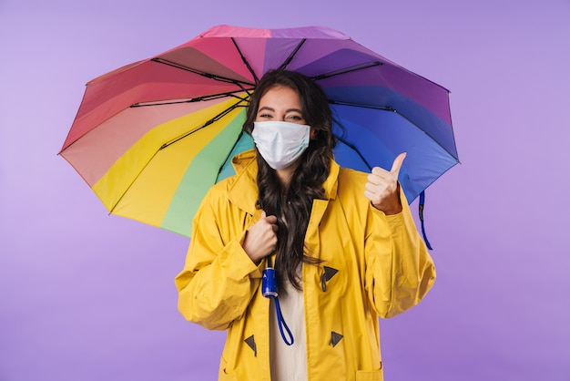 donna ottimista positiva in impermeabile giallo in posa isolata sul muro viola che tiene l'ombrello che indossa una maschera medica che mostra i pollici in su.