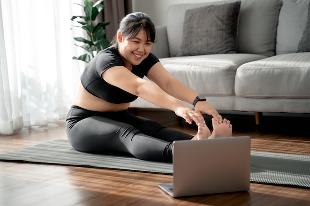 donna paffuta seduta sul pavimento in soggiorno pratica lezione di yoga online con il computer