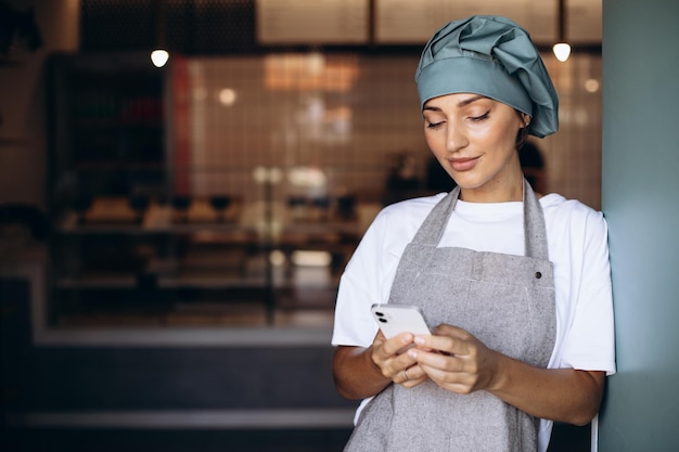 Donna panettiere in grembiule in piedi con il telefono vicino alle porte della caffetteria