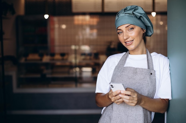 Donna panettiere in grembiule in piedi con il telefono vicino alle porte della caffetteria