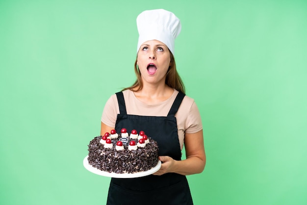 Donna pasticcera di mezza età che tiene una grande torta su sfondo isolato guardando in alto e con espressione sorpresa