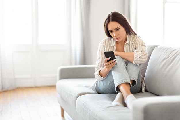 Donna pensierosa con uno smartphone sul divano a casa