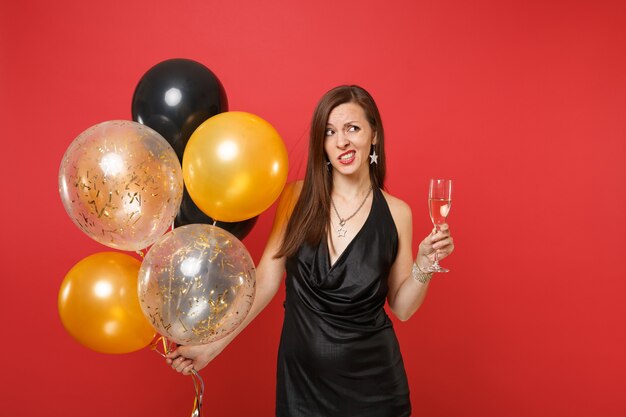 Donna pensierosa preoccupata in abito nero che celebra, tenendo in mano un bicchiere di champagne e mongolfiere isolate su sfondo rosso. San Valentino, felice anno nuovo, concetto di festa di compleanno mockup.