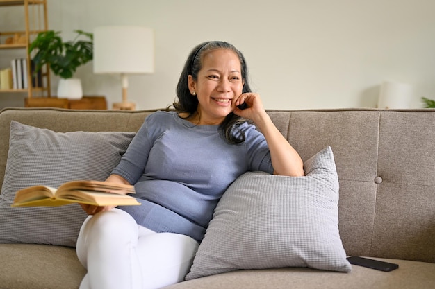 Donna pensionata asiatica sorridente e felice che si rilassa su un divano nel suo salotto