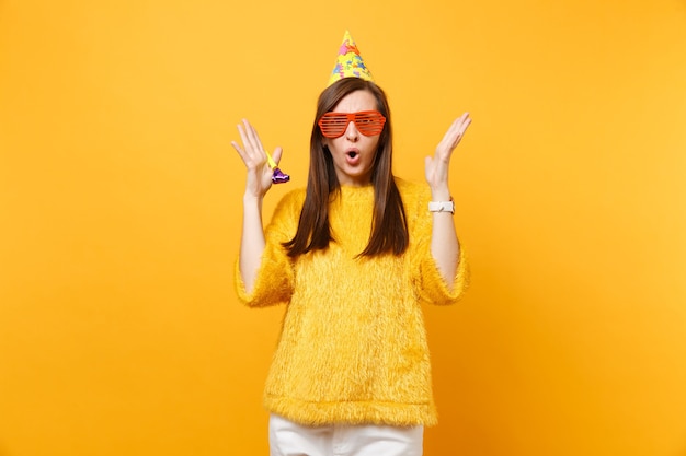 Donna perplessa scioccata con occhiali divertenti arancioni, cappello di compleanno con pipa che allarga le mani, celebrando isolato su sfondo giallo. Persone sincere emozioni, concetto di stile di vita. Zona pubblicità.
