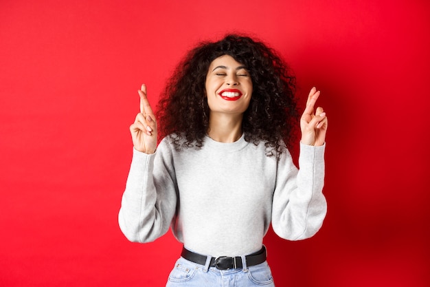 Donna piena di speranza e positiva con labbra rosse e capelli ricci, incrociare le dita per buona fortuna e esprimere desideri, pregando per il sogno che si avvera, sorridente eccitato, sfondo rosso.