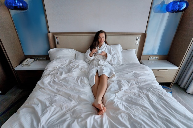 Donna pigra in accappatoio sdraiato sul letto e godersi la colazione del buongiorno con una tazza di caffè aromatica durante il relax nell'accogliente camera da letto comfort nella camera d'albergo
