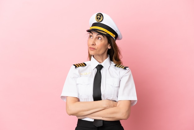 Donna pilota di mezza età dell'aeroplano isolata su sfondo rosa che guarda in alto mentre sorride