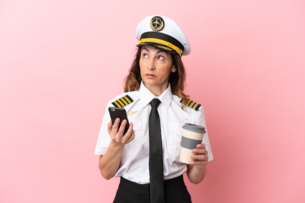 Donna pilota di mezza età dell'aeroplano isolata su sfondo rosa che tiene il caffè da portare via e un cellulare mentre pensa a qualcosa