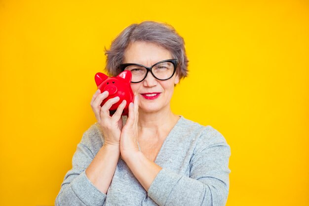 Donna più anziana caucasica che tiene banca piggy che posa felicemente per la macchina fotografica su fondo giallo