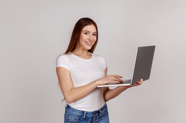 Donna positiva felice che tiene laptop, guarda la fotocamera, naviga in Internet, lavora come studente o libero professionista, usa il computer, indossa una maglietta bianca. Studio indoor girato isolato su sfondo grigio.