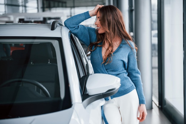 Donna positiva in camicia blu appoggiata su un'auto nuova di zecca Nel salone dell'auto