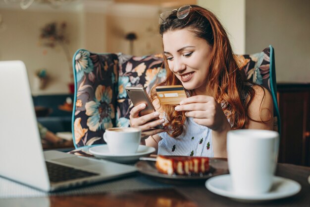 Donna positiva per il corpo con i capelli rossi che tiene una carta di credito e uno smartphone che fa transazione online mentre era seduto in una caffetteria con un laptop sulla sua scrivania.