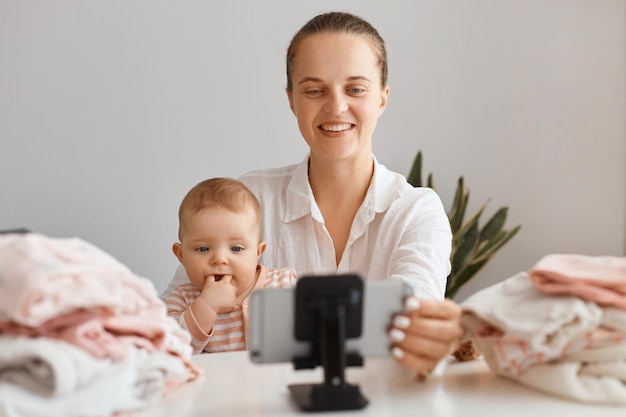 Donna positiva seduta al tavolo con un bambino piccolo e che registra video per il suo vlog, realizza contenuti con sua figlia, guardando sorridendo alla fotocamera dello smartphone, livestreaming.