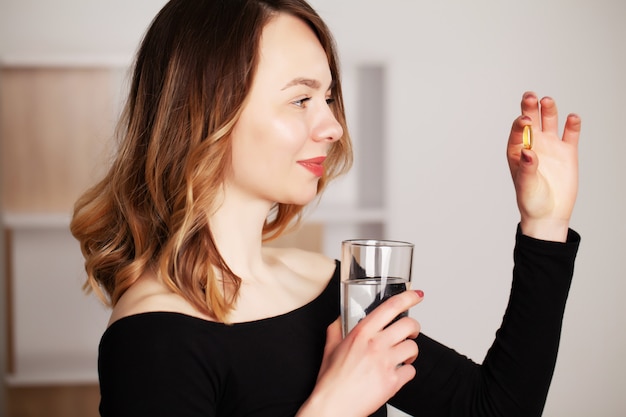 Donna positiva sorridente felice che mangia la pillola e che tiene il bicchiere d'acqua nella mano, nella sua casa