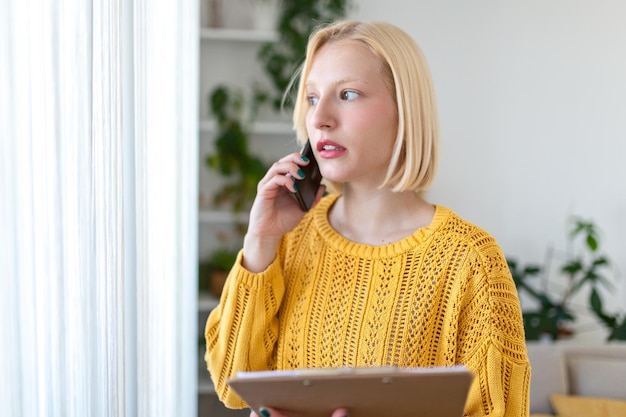 Donna premurosa che tiene carta mentre parla al telefono cellulare. La femmina sta guardando attraverso la finestra. Lei è a casa.