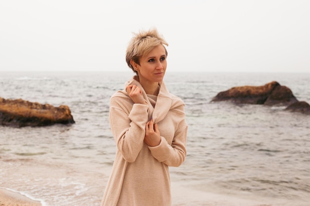 Donna premurosa hipster con taglio di capelli corto che indossa un abito beige vicino al mare in autunno