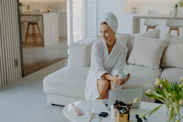 Donna premurosa rilassata vestita in accappatoio e asciugamano avvolto sulla testa si siede sul divano con una tazza di tè caldo