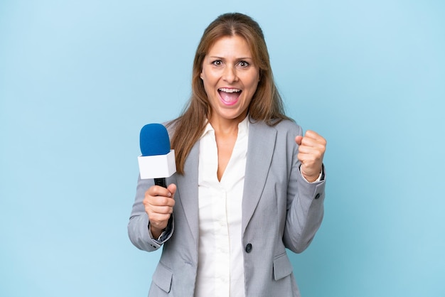 Donna presentatrice televisiva di mezza età su sfondo blu isolato che celebra una vittoria nella posizione del vincitore