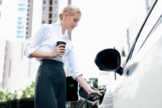 Donna progressista con caffè mentre carica un'auto EV con edifici residenziali