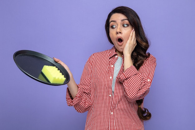 Donna pulita piuttosto caucasica sorpresa che tiene e guarda la spugna sul piatto e le mette la mano sul viso