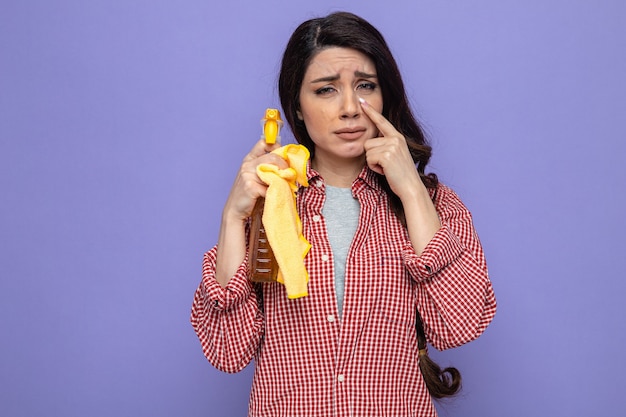 Donna pulita piuttosto caucasica triste che tiene il dito vicino all'occhio e tiene in mano panni per la pulizia e detergente spray