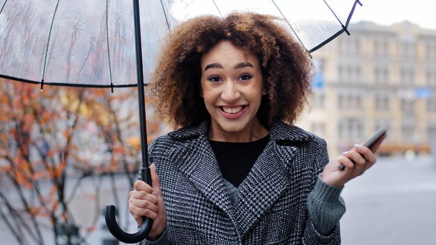 Donna ragazza afroamericana con ombrello trasparente si trova in città sotto la pioggia guardando video divertenti