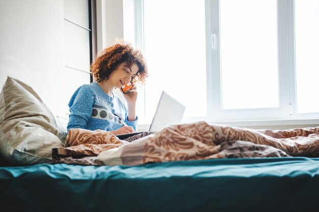 Donna riccia che parla al telefono direttamente dal letto notando le lezioni online dei compiti